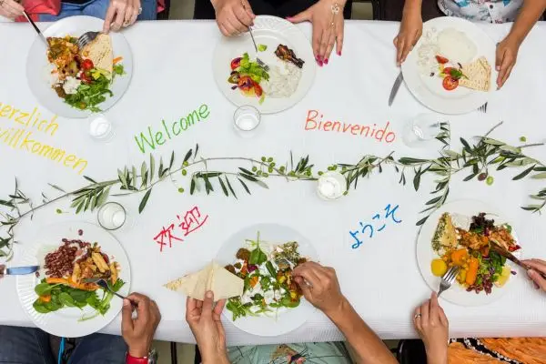 A diverse, multilingual dinner table with 'Welcome' greetings in various languages, symbolizing culturally adapted food localization and global agribusiness expansion with Intervoices Global.